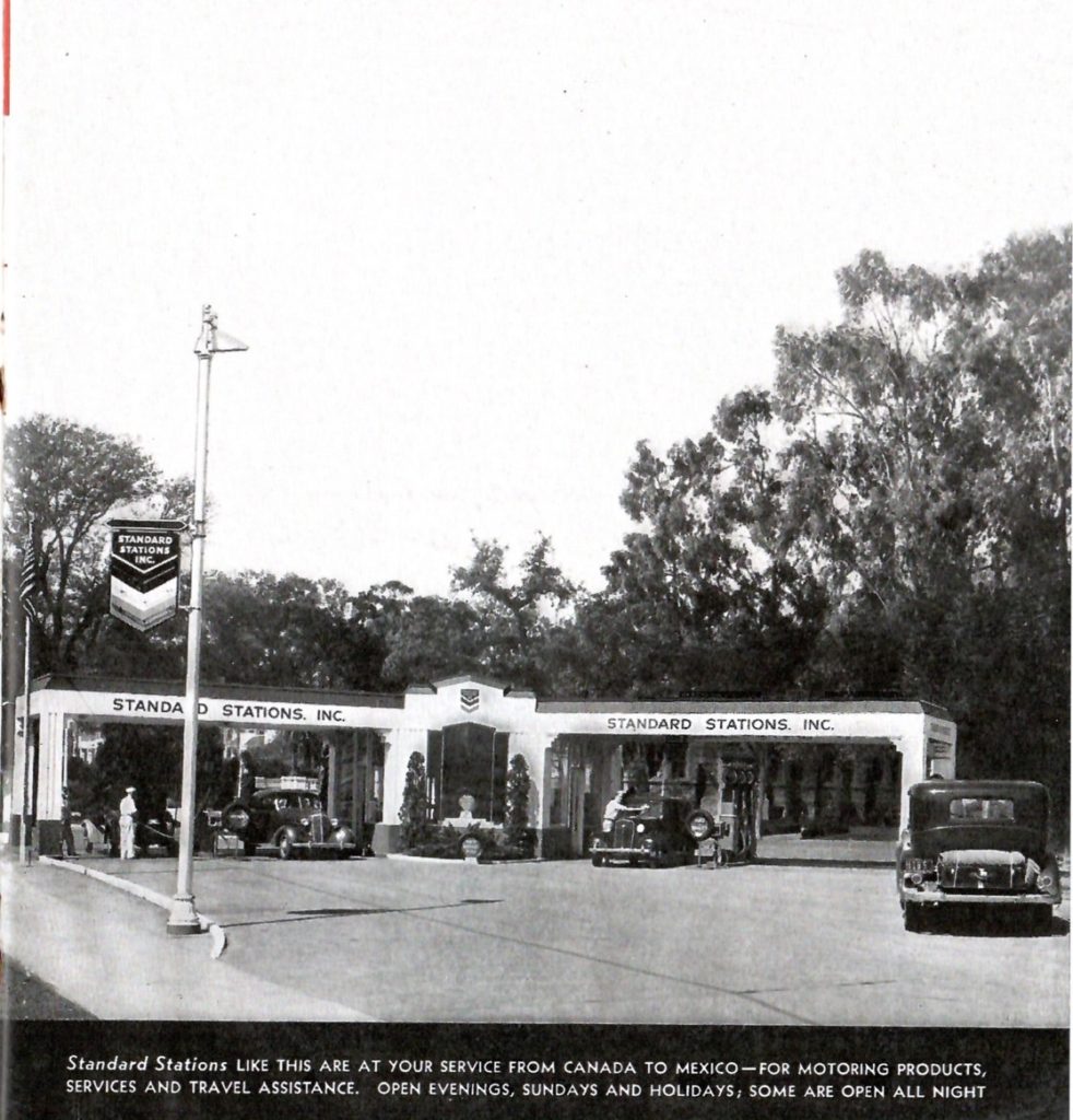 A black and white photo of a service station in 1936. It is run by Standard Stations. It has two vintage cars are getting gasoline, and being tended to buy service station men wearing hats.